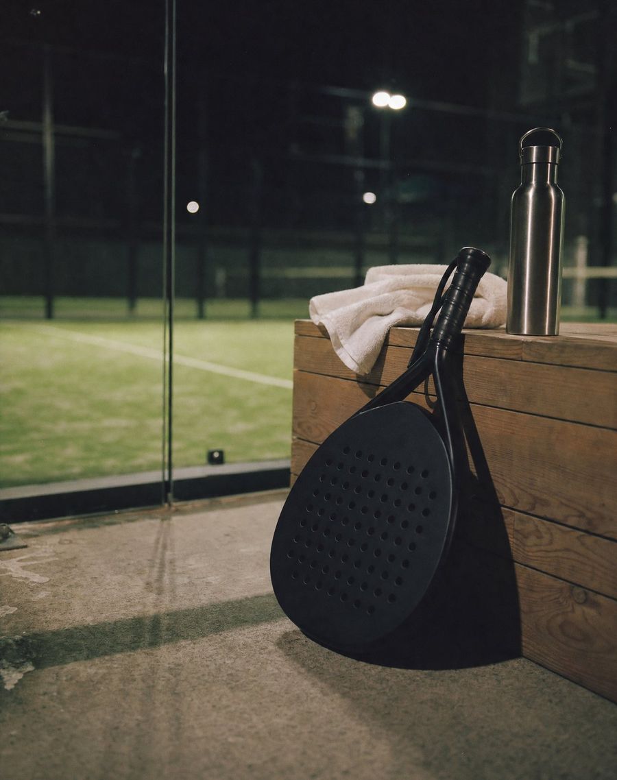 A padel racquet leaning on a courtside timber bench beside a towel and a water bottle, the court lit behind.